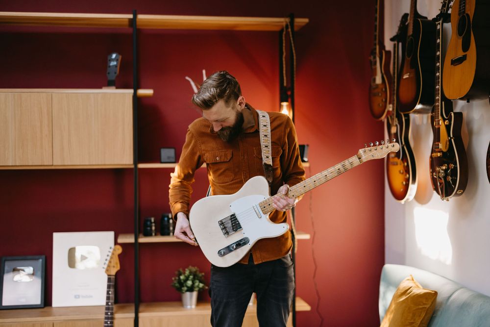 Musician in a brown jacket playing white telecaster guitar in music room with wall-mounted guitars.