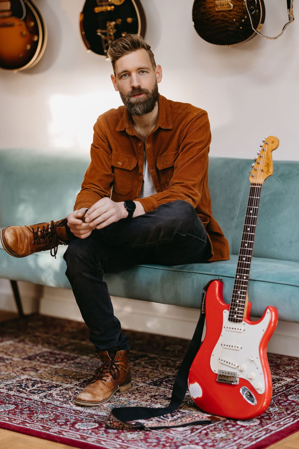 Musician seated on blue couch with red Stratocaster guitar against wall of hanging instruments.