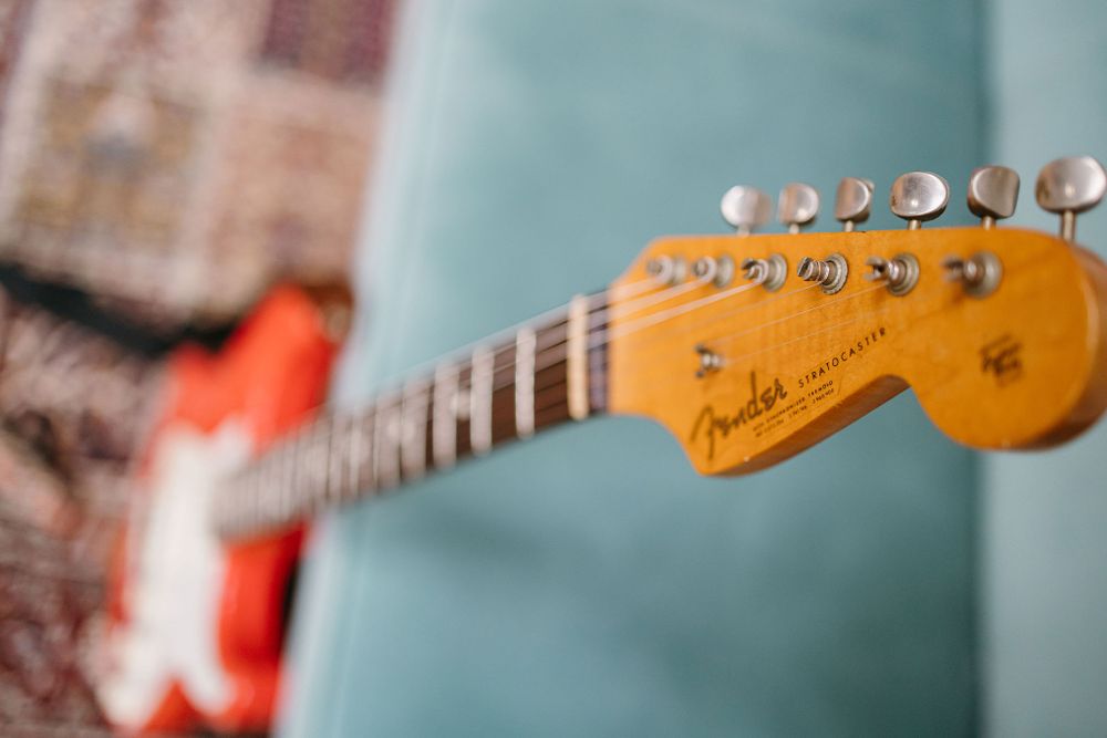 Close-up view of a Fender guitar headstock with chrome tuning pegs against a blurred teal background.