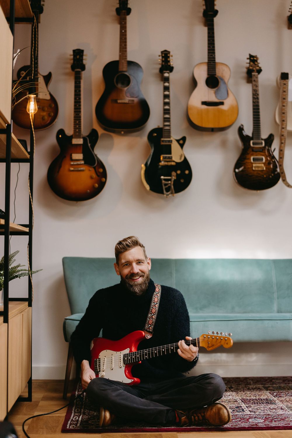 Musician sitting cross-legged on floor playing electric guitar with collection of guitars mounted on wall behind.