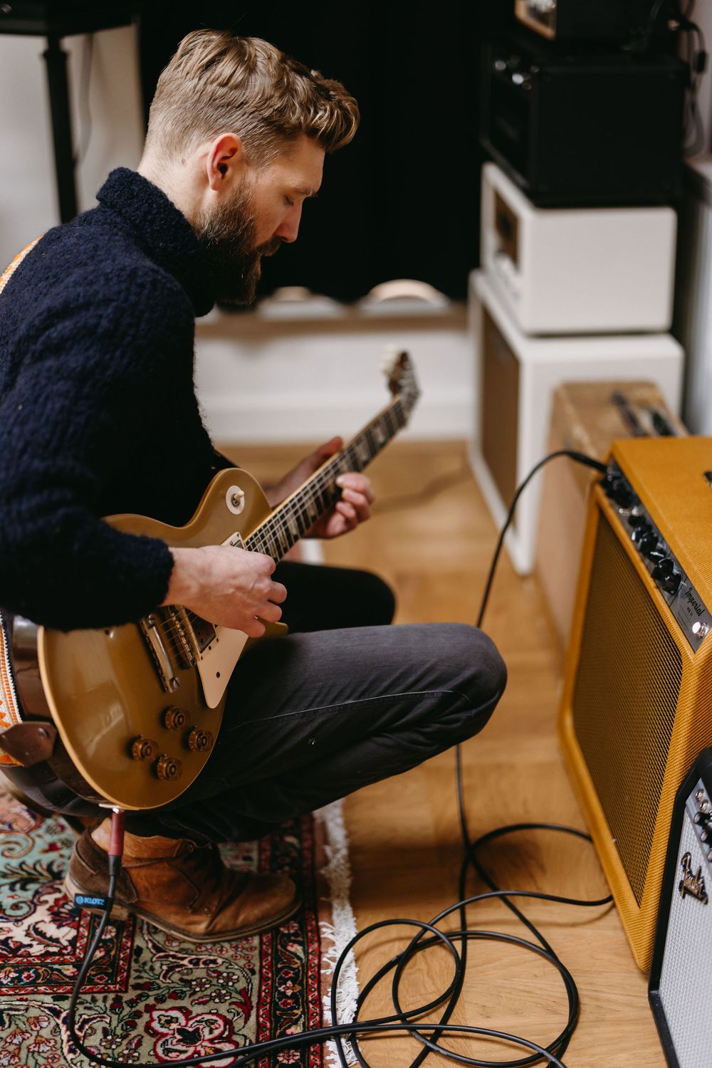 Musician playing gold Les Paul guitar while sitting beside vintage amplifier on decorative rug.