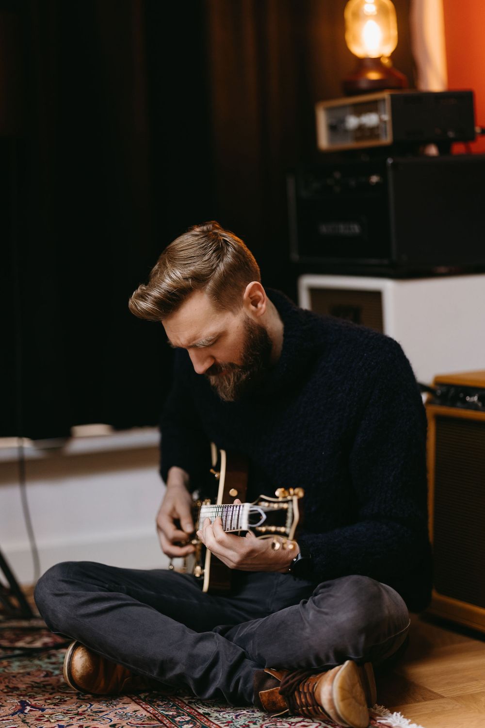 Guitar player sitting cross-legged on oriental rug practicing acoustic guitar in dimly lit studio.