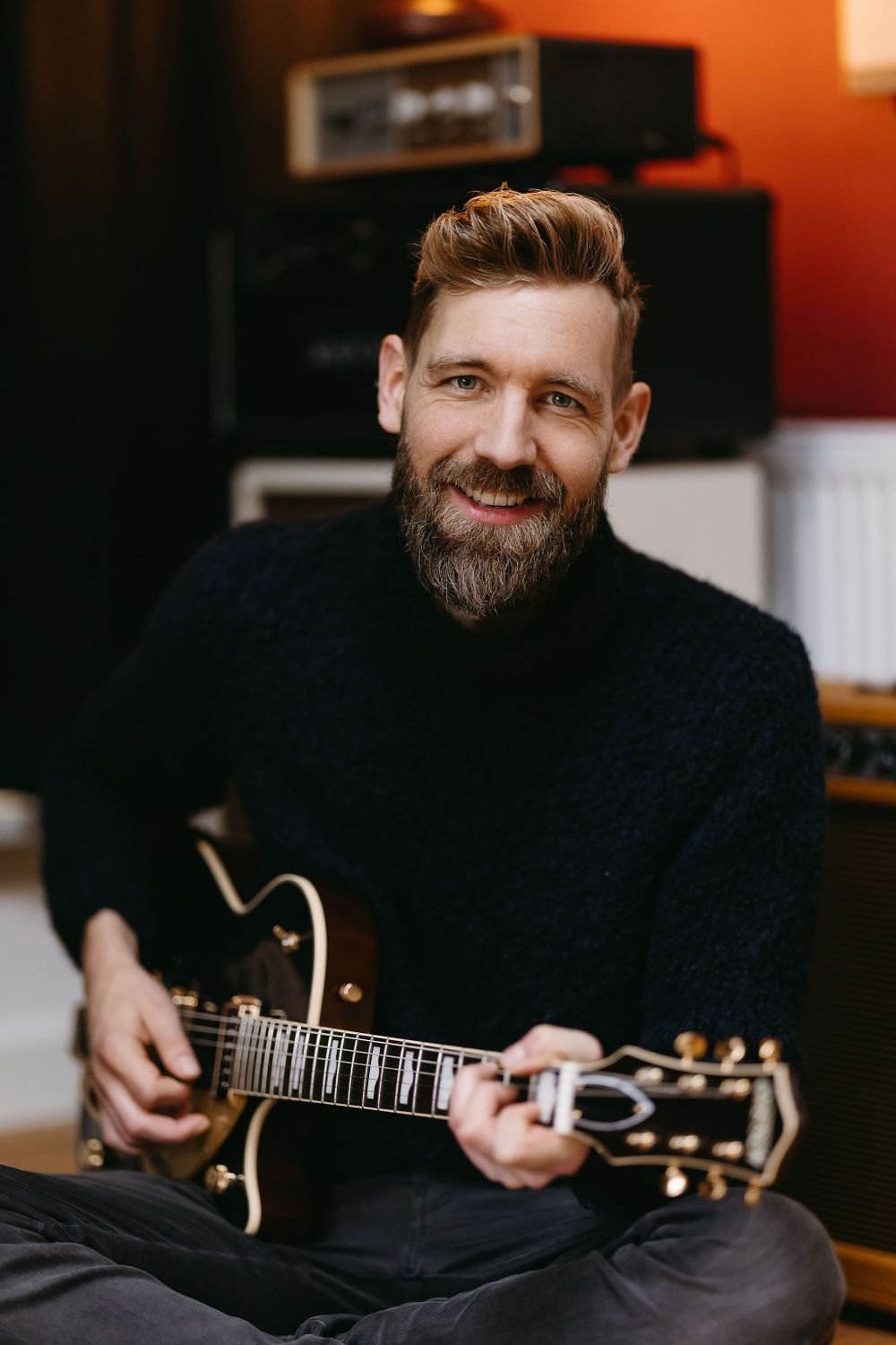 Musician in black sweater playing guitar in recording studio setting.