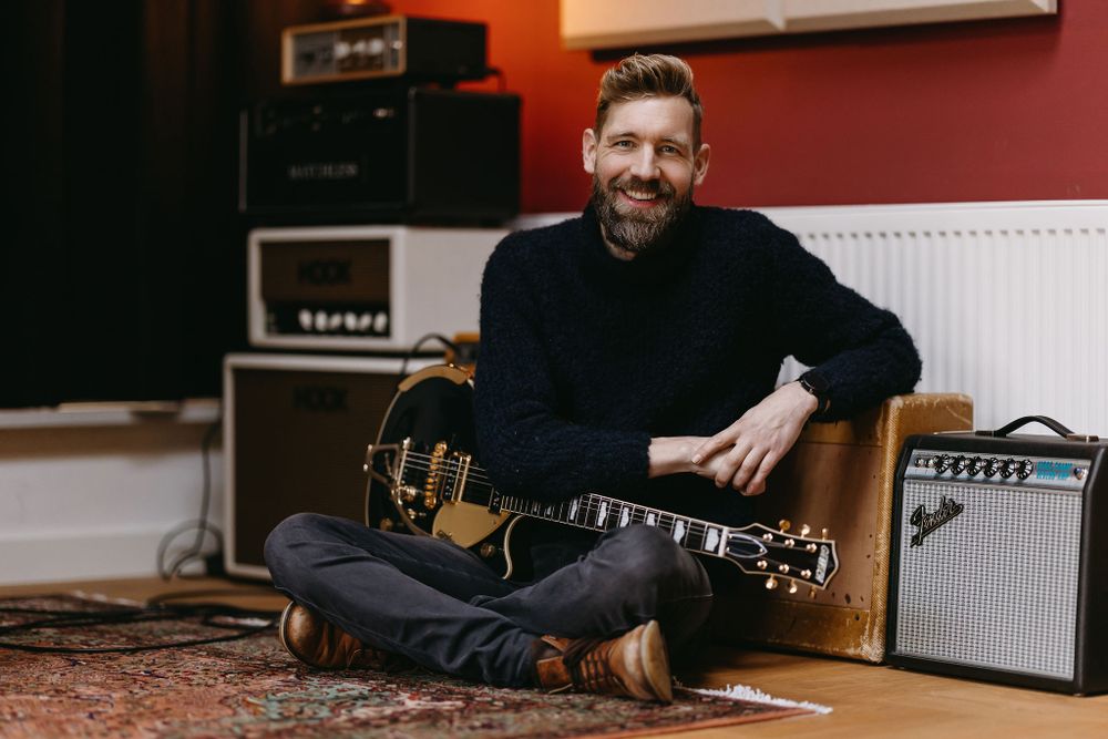 Musician in black sweater sitting with acoustic guitar in recording studio environment.
