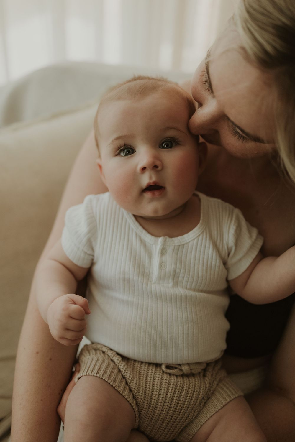 A mother tenderly kisses her baby's head as the infant in a white ribbed onesie looks curiously at the camera.