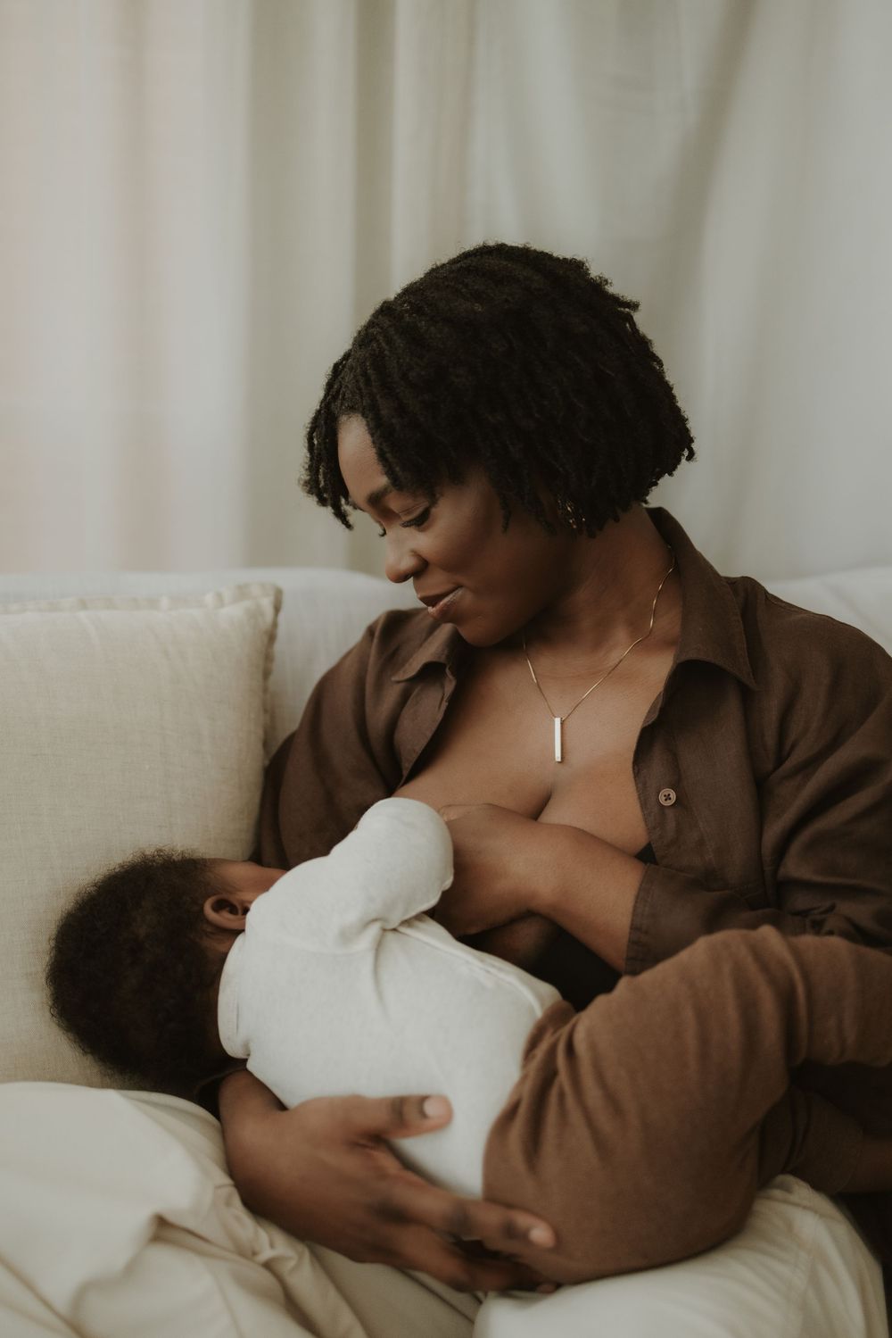 A Black mother breastfeeding her newborn baby while sitting on a white sofa, wearing a brown shirt.