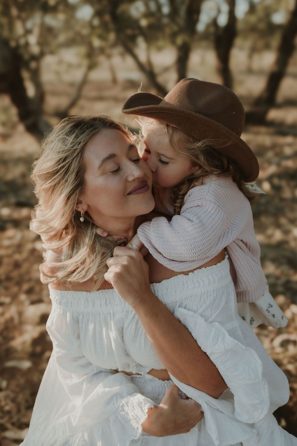 A mother and child share tender moments in a sun-dappled outdoor setting wearing white clothing.