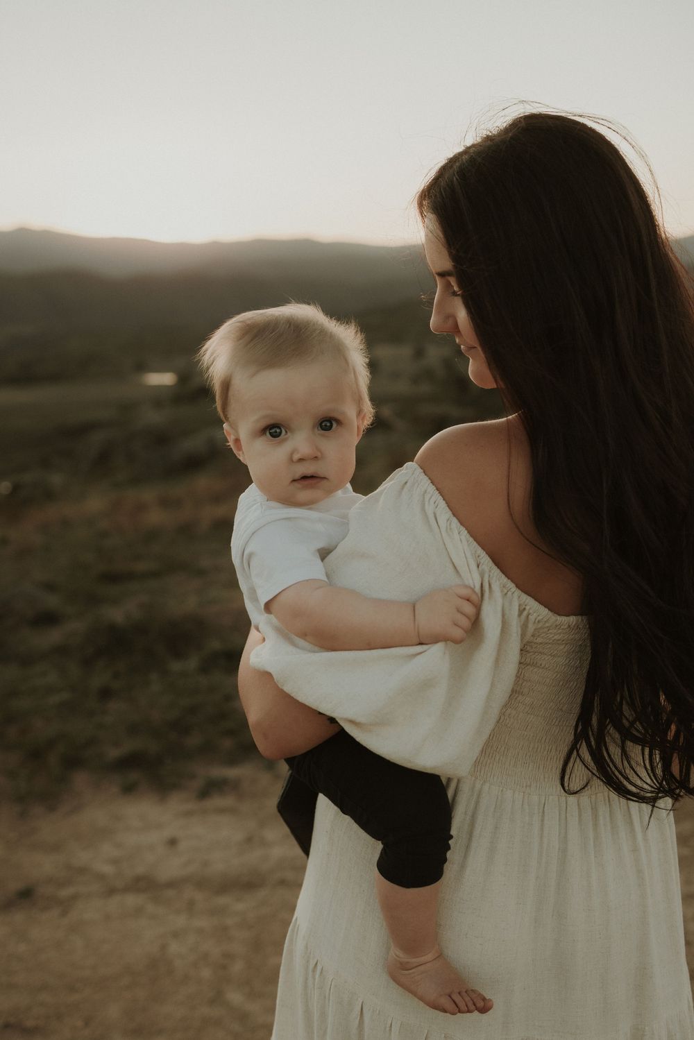 A parent in a cream dress holds a baby in white against a moody outdoor landscape at sunset.