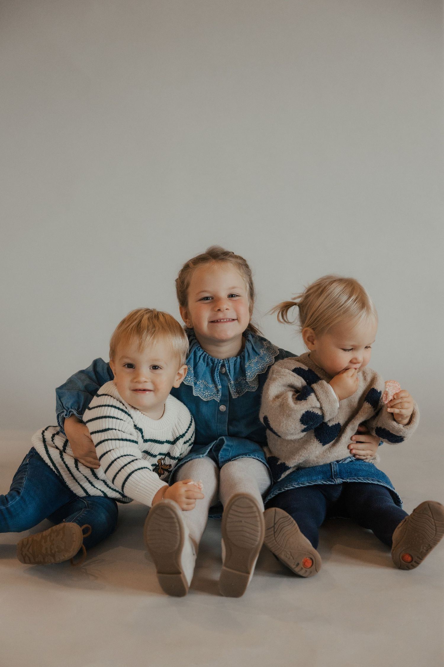 Three young children sitting together on a light gray backdrop wearing casual clothing.