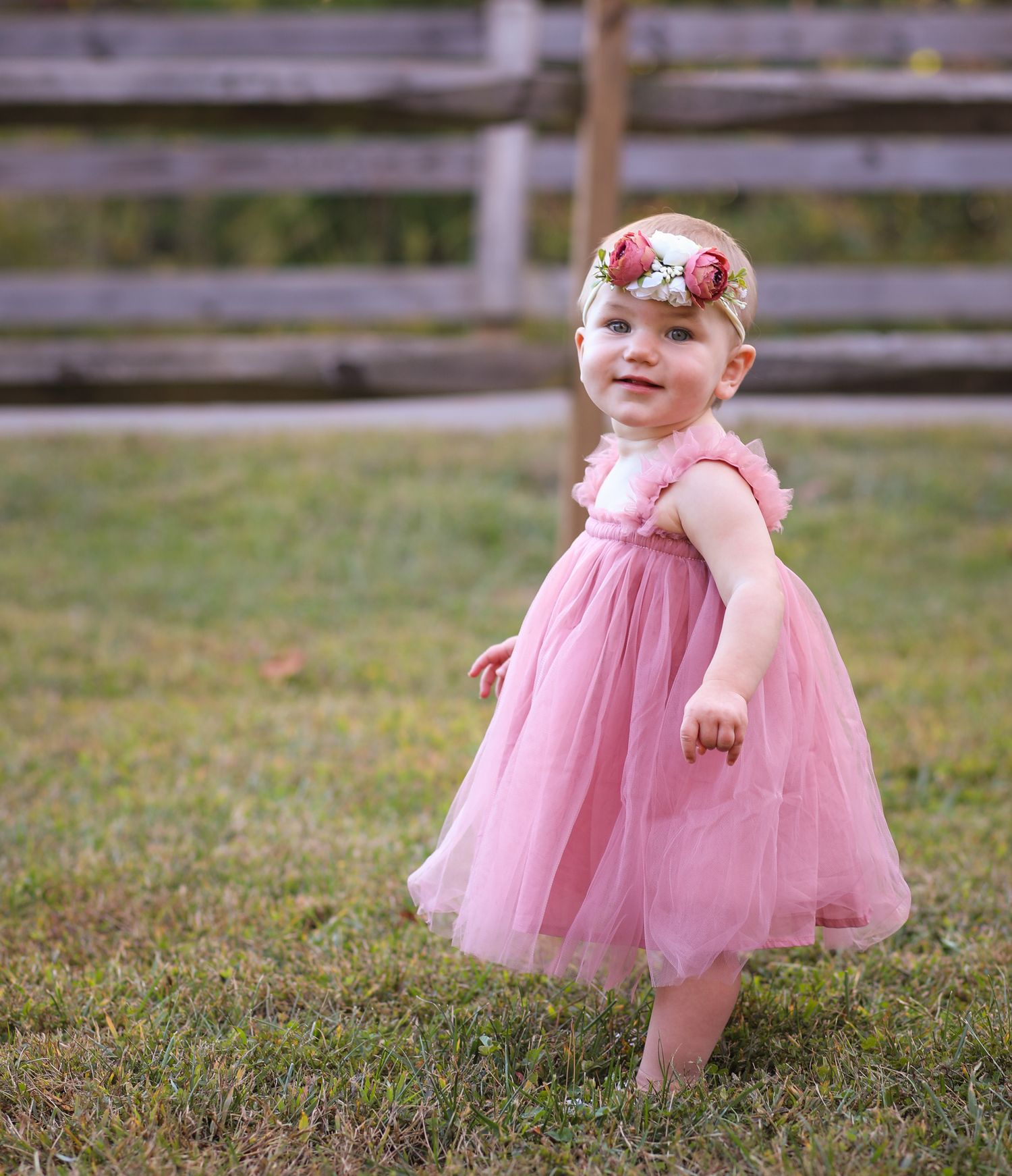 Smiling toddler girl in pink tutu dress and floral headband standing on green grass outdoors.