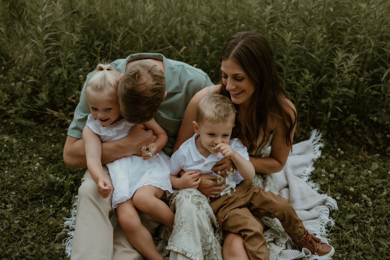 A family sits together on a white blanket outdoors surrounded by greenery on a summer day.