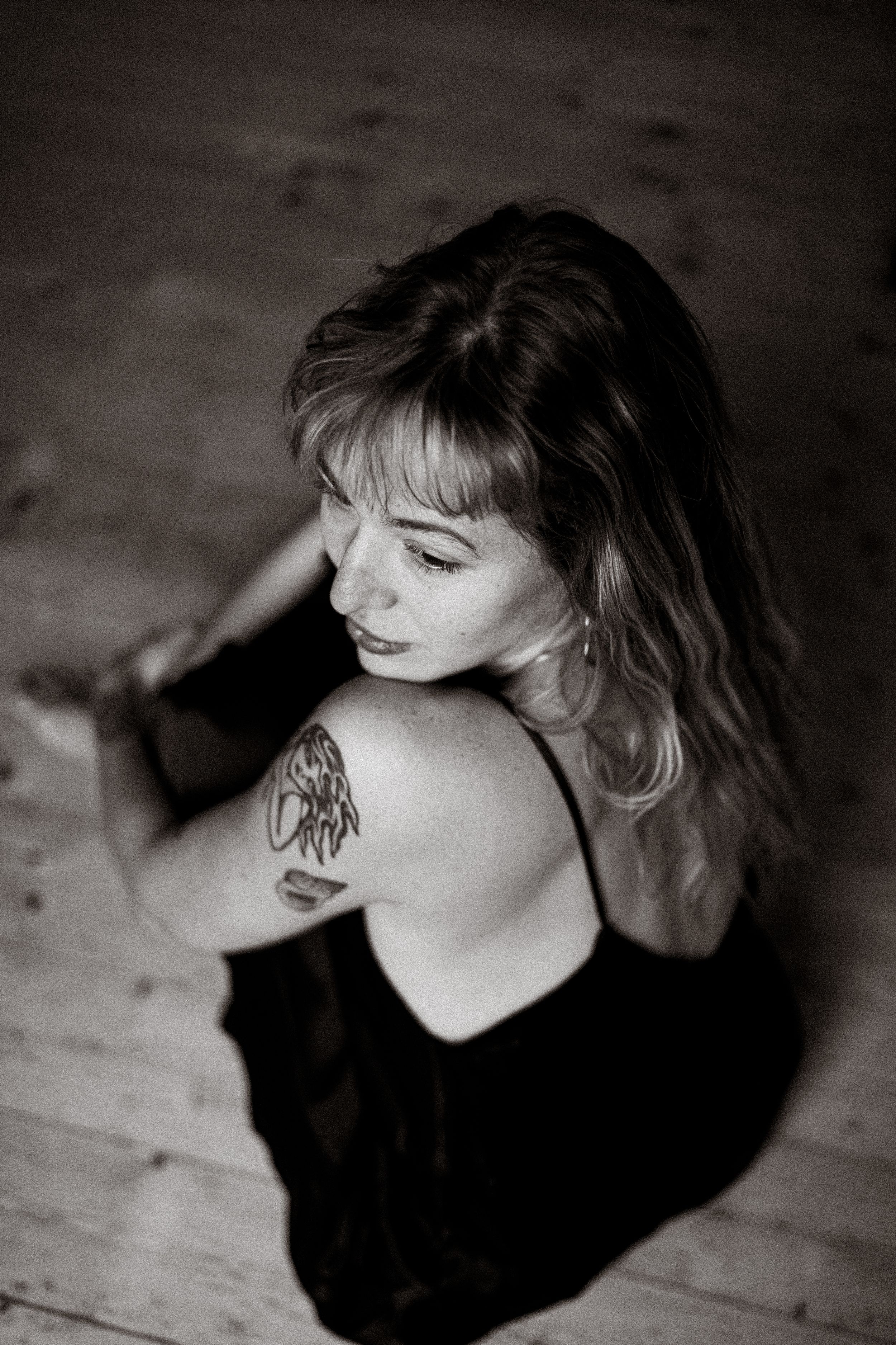 Black and white photo of a woman with wavy hair and shoulder tattoo, seated on wooden floor, wearing black dress.