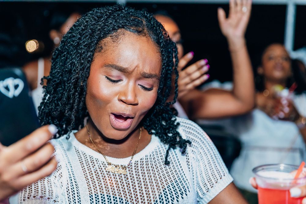 Woman with braids singing passionately in white knit top at party.