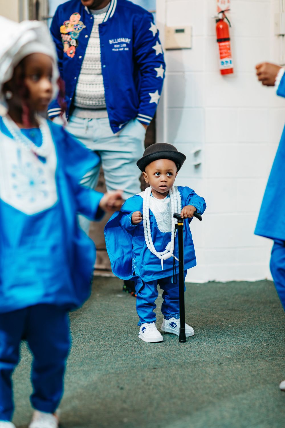 A child in blue attire wearing a hat poses with a cane against a white wall at a special event.