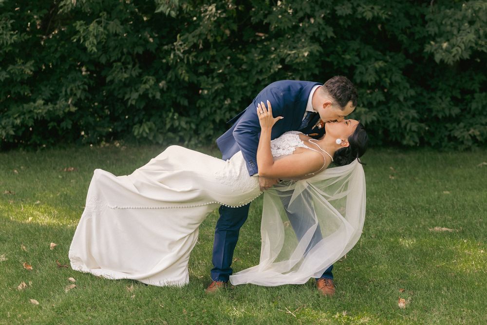 Bride and groom sharing a romantic dip kiss on green grass with lush trees in the background at outdoor wedding.