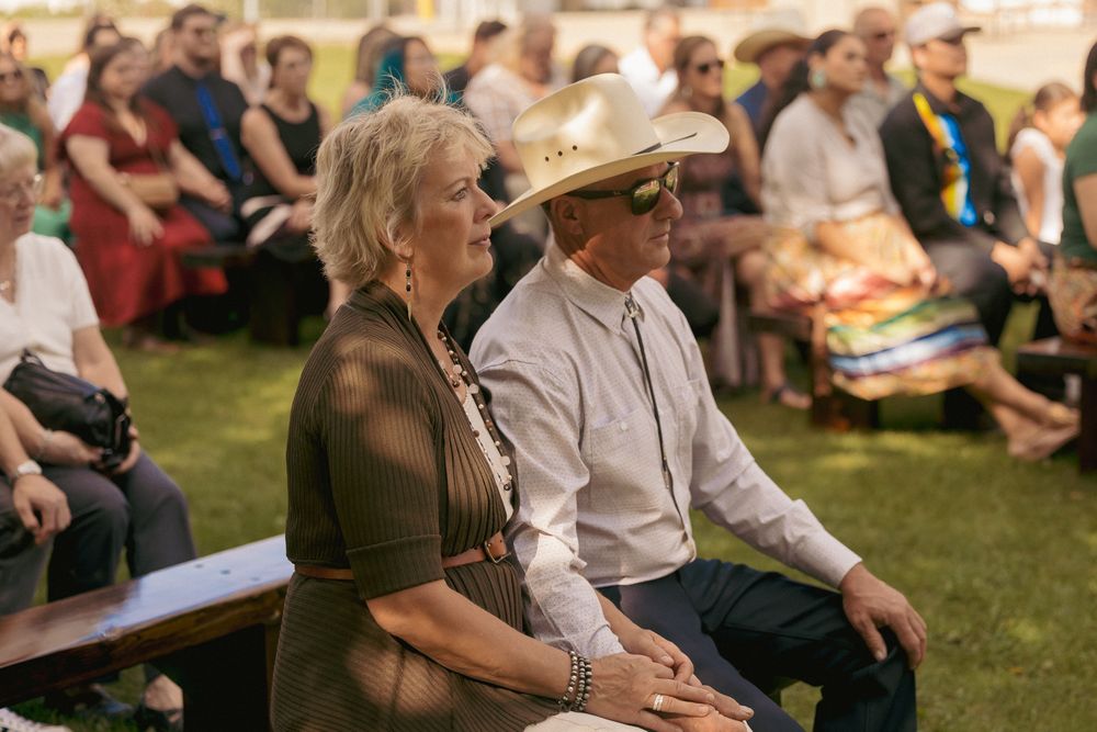 Couple in western attire sitting on bench at outdoor event, man wearing cowboy hat, surrounded by crowd on grass.