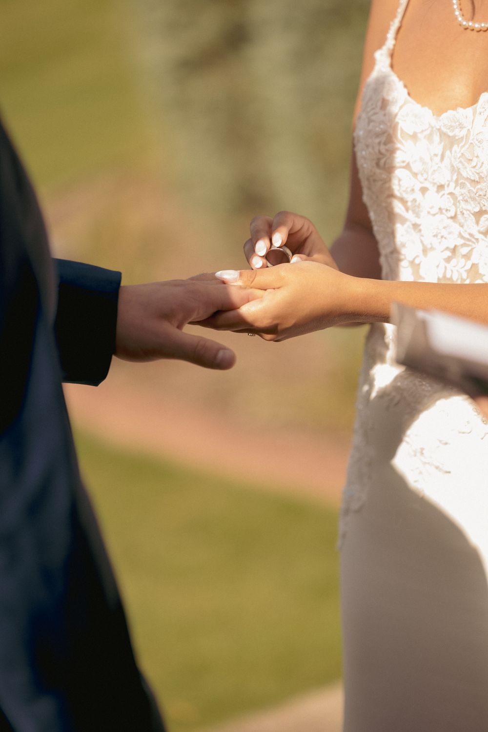 Interracial couple exchanging wedding rings during outdoor ceremony, groom in navy suit placing ring on brides finger.