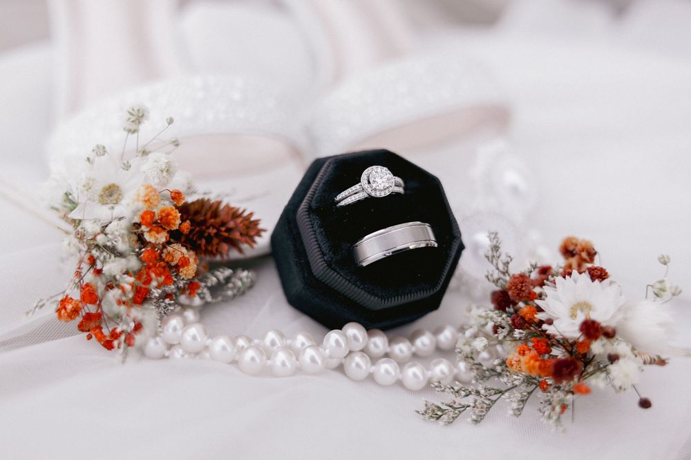 Diamond engagement ring and wedding band displayed in black velvet box with dried flowers and pearl necklace.