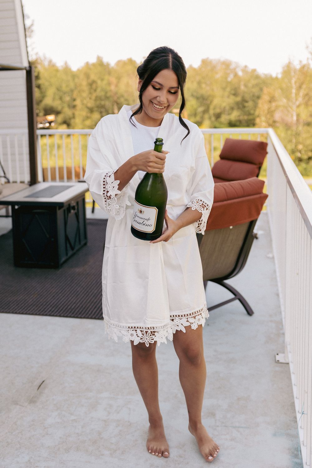 Smiling woman in white lace robe opening champagne bottle on outdoor balcony with green trees in background.