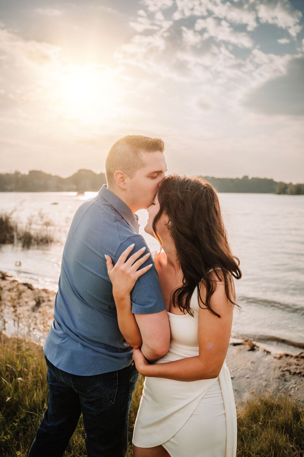 A romantic sunset beach engagement photo session featuring a couple embracing with water and soft clouds in background.