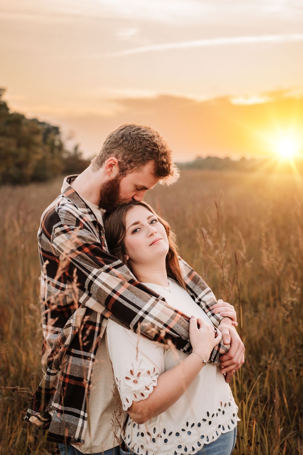Romantic couple embraces in golden wheat field at sunset while wearing casual plaid and white clothing.