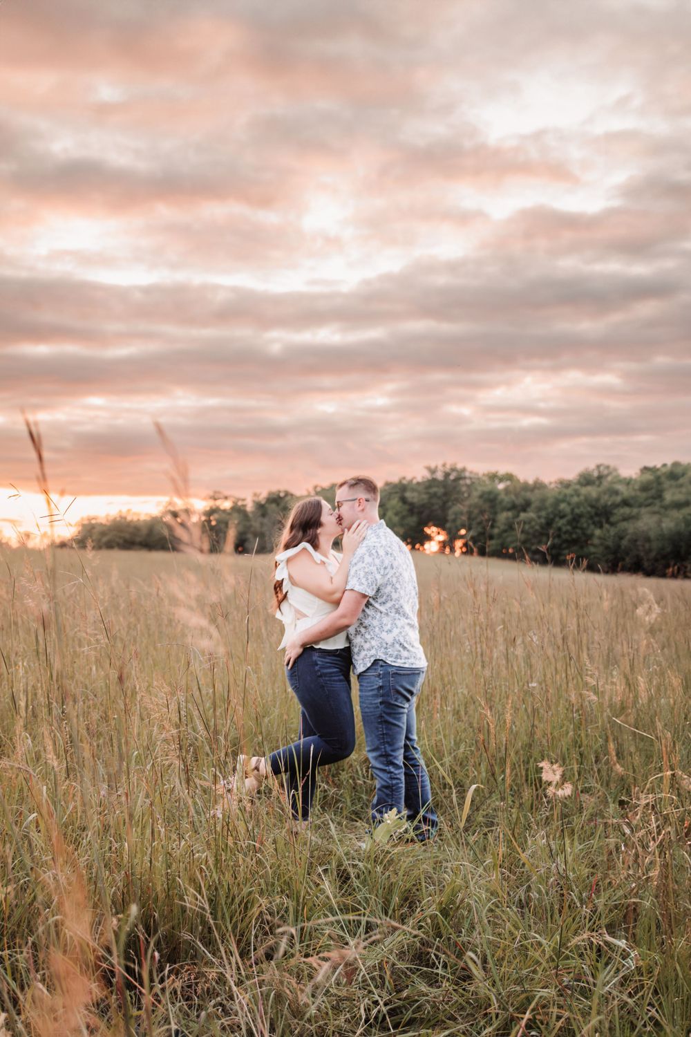 Romantic couple embracing in tall grass field during pink sunset.
