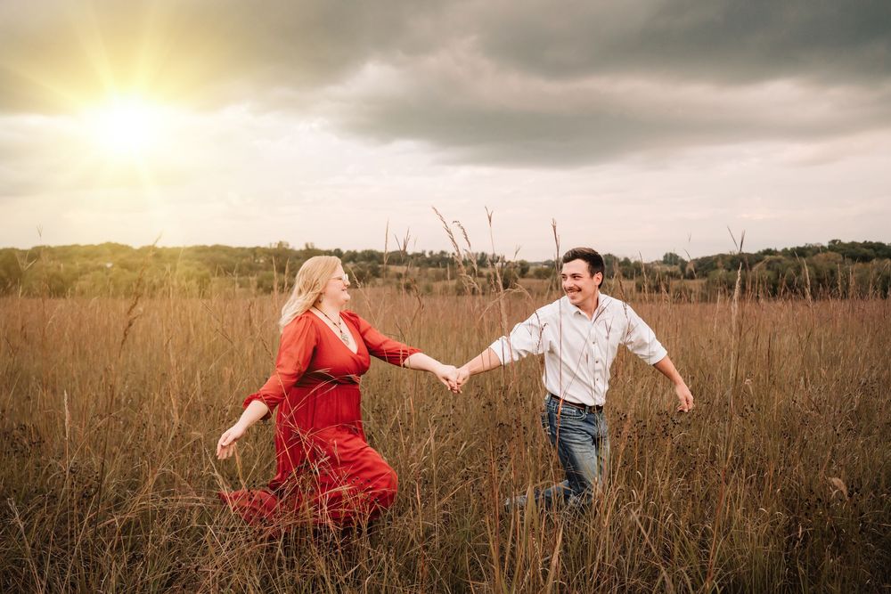 A couple in a field holds hands and dances playfully through tall golden grass at sunset.