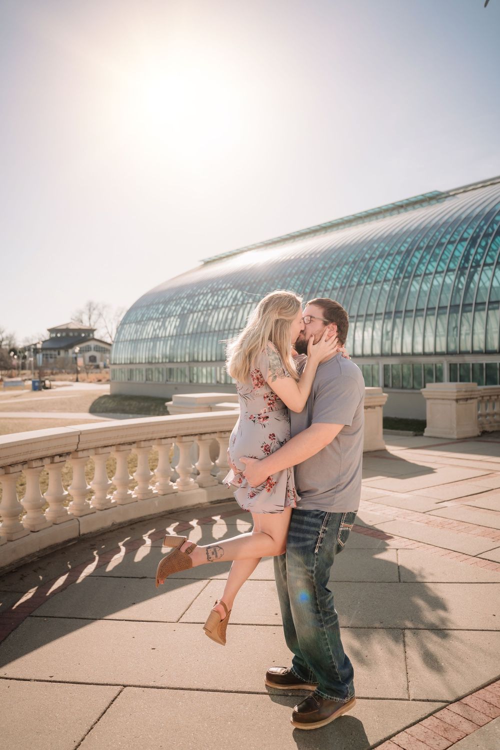 Couple sharing a romantic kiss outdoors near a glass greenhouse on a sunny day.