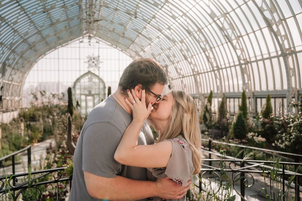 Couple sharing a romantic kiss inside a sunlit greenhouse with arched glass ceiling and lush greenery.