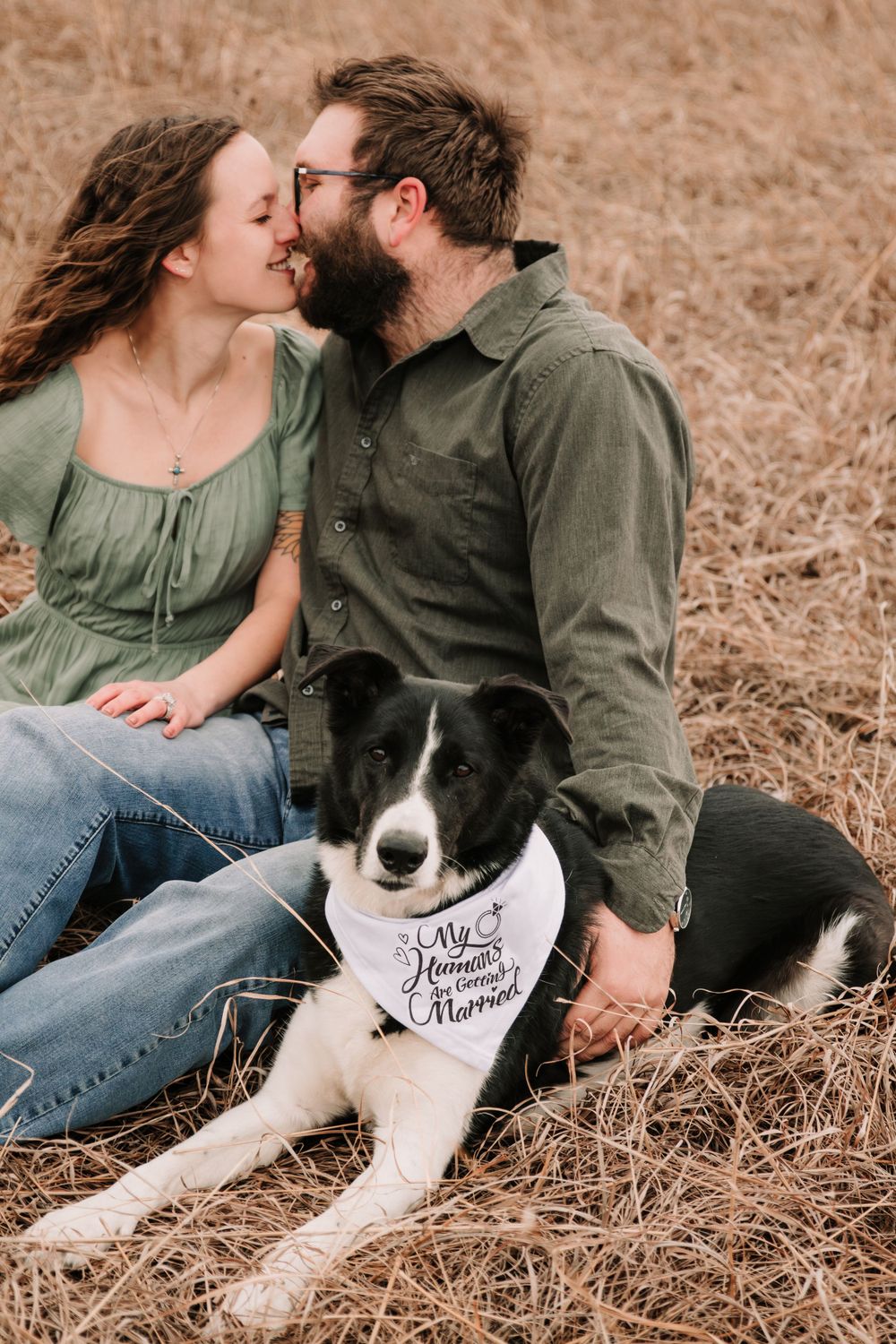 Engagement photo featuring a couple and their border collie wearing a bandana in a field.