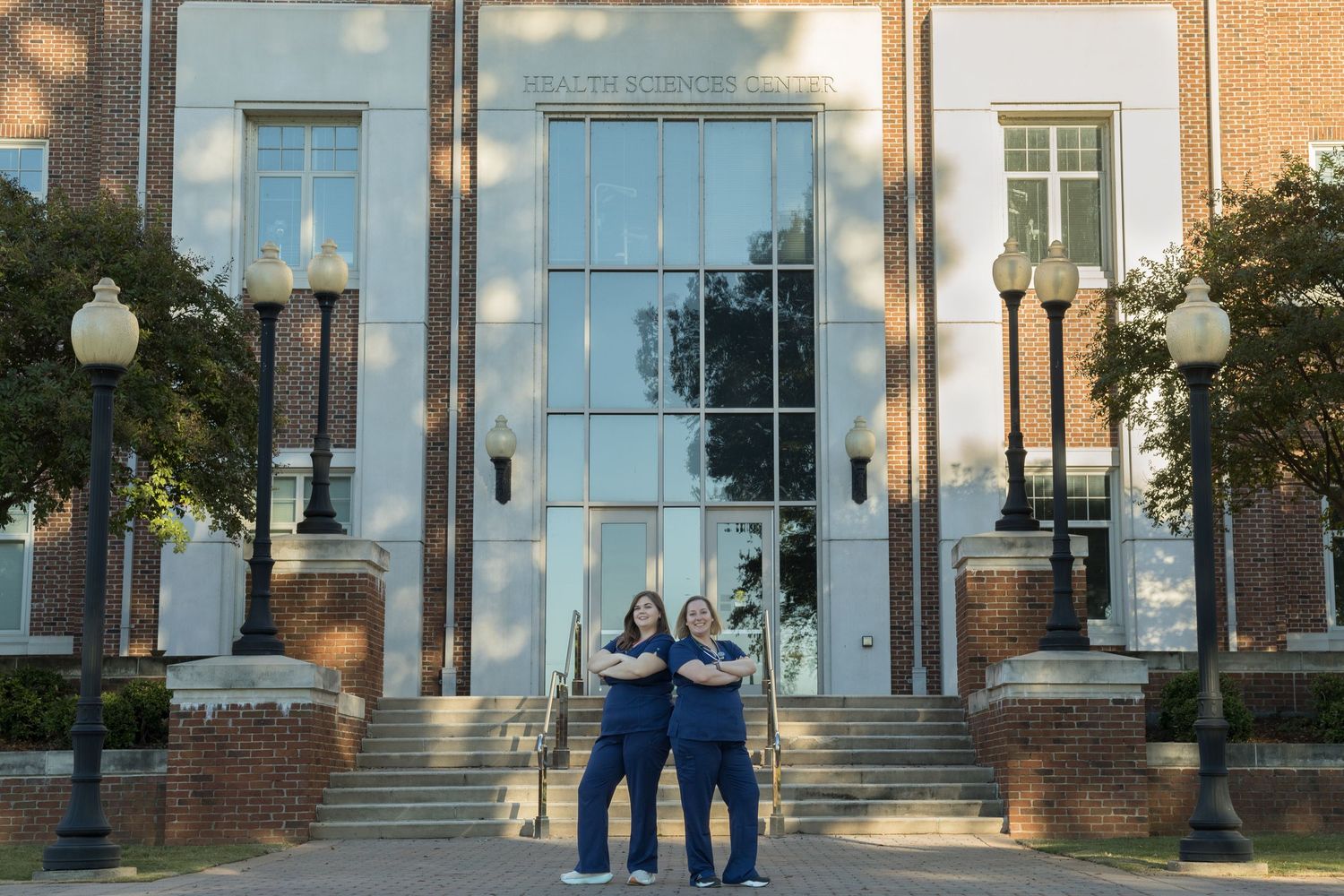 Two nursing students stand in front of the nursing building at their college.