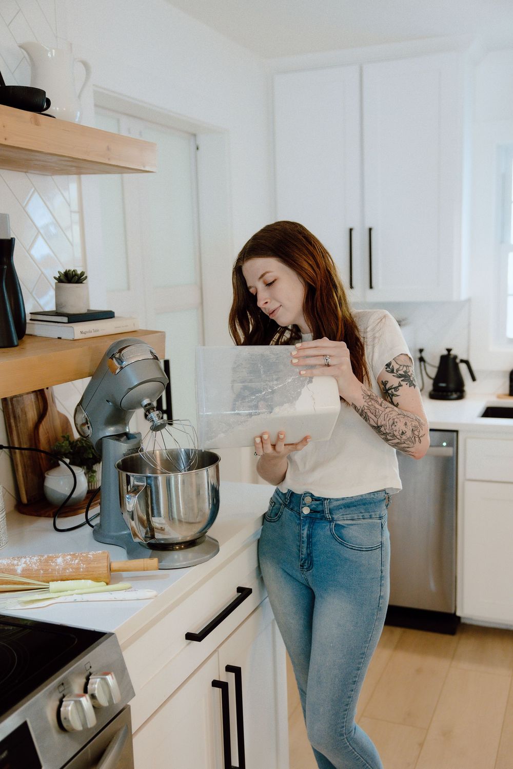 Baker in white sweater and jeans using a stand mixer in a bright modern kitchen.