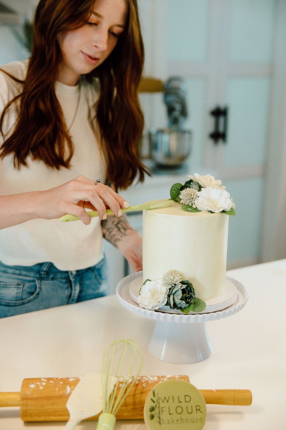 White cake being decorated with fresh flowers on a marble countertop.