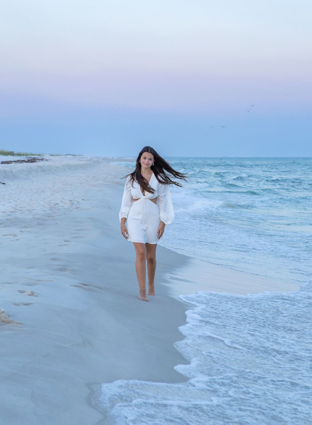 A girl in white walks down the beach in Perdido Key, Floroida at sunset with her hair blowing for senior portraits