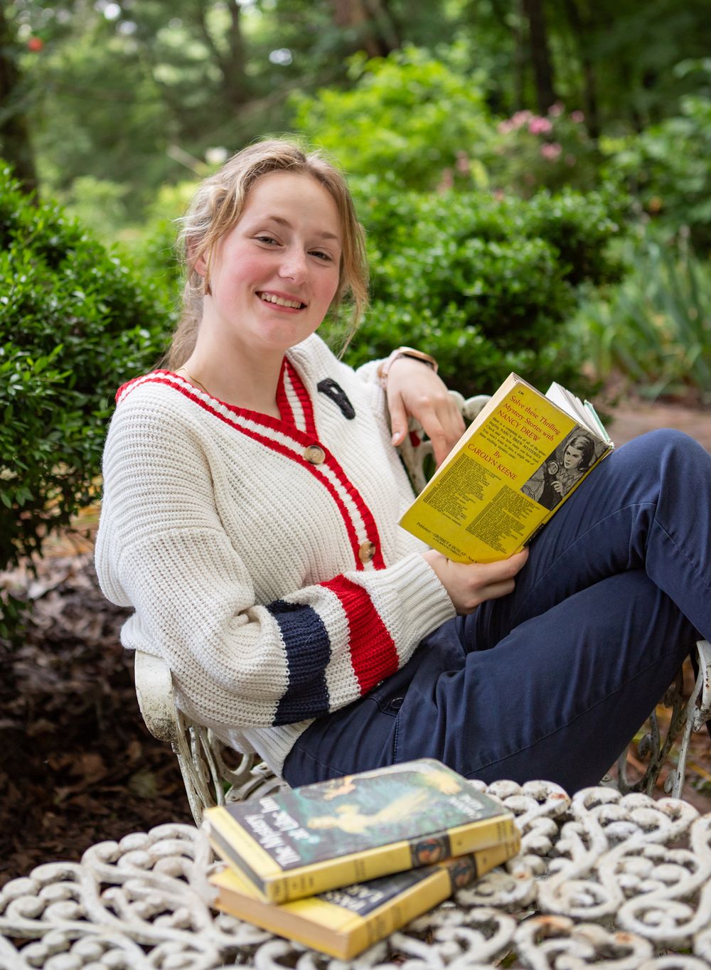 A high school senior girl wears a preppy sweater and reads in a garden for her senior portrait session in Pensacola