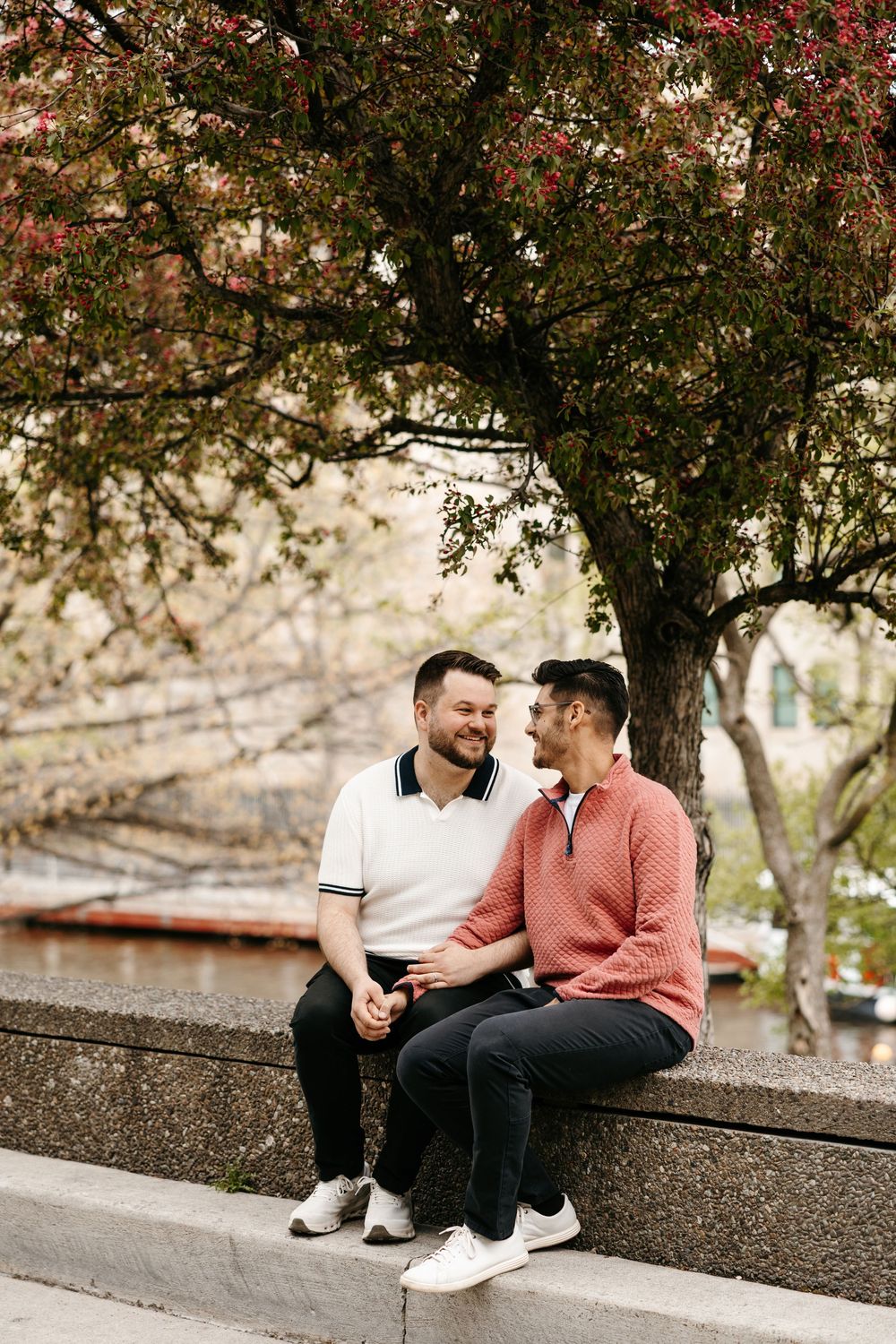 Two men sitting close together on a stone ledge under a flowering tree, smiling at each other in an outdoor park setting.