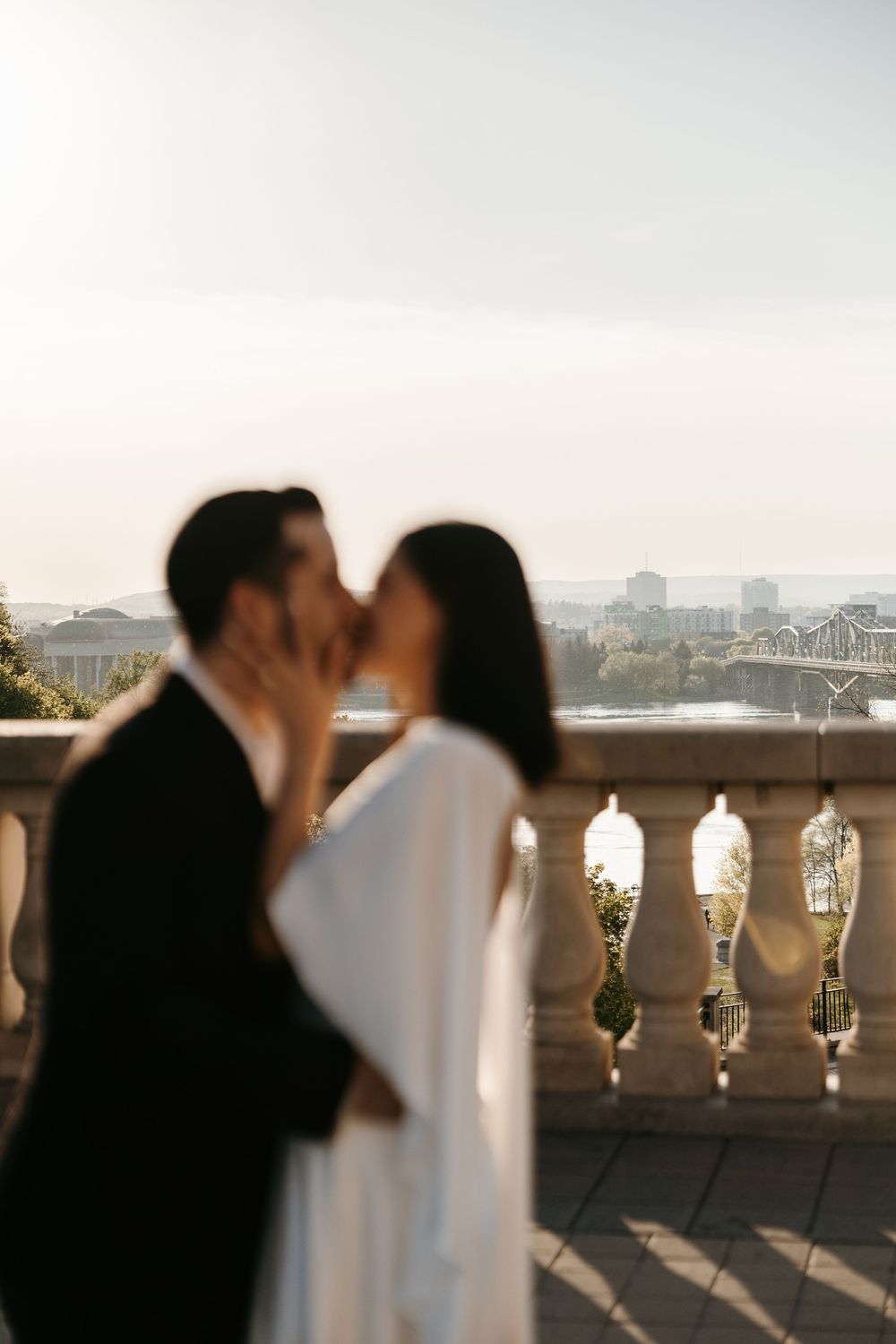 A couple shares a romantic kiss on a sunlit balcony with city views, the bride in white and groom in black attire.