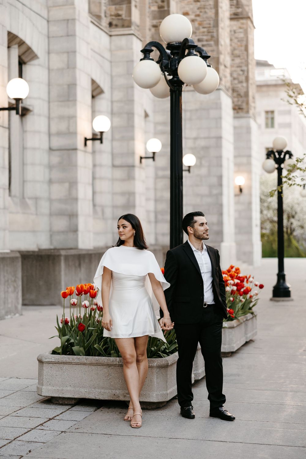 Couple posing near ornate street lamp and red tulips outside historic building, woman in white dress, man in black suit.