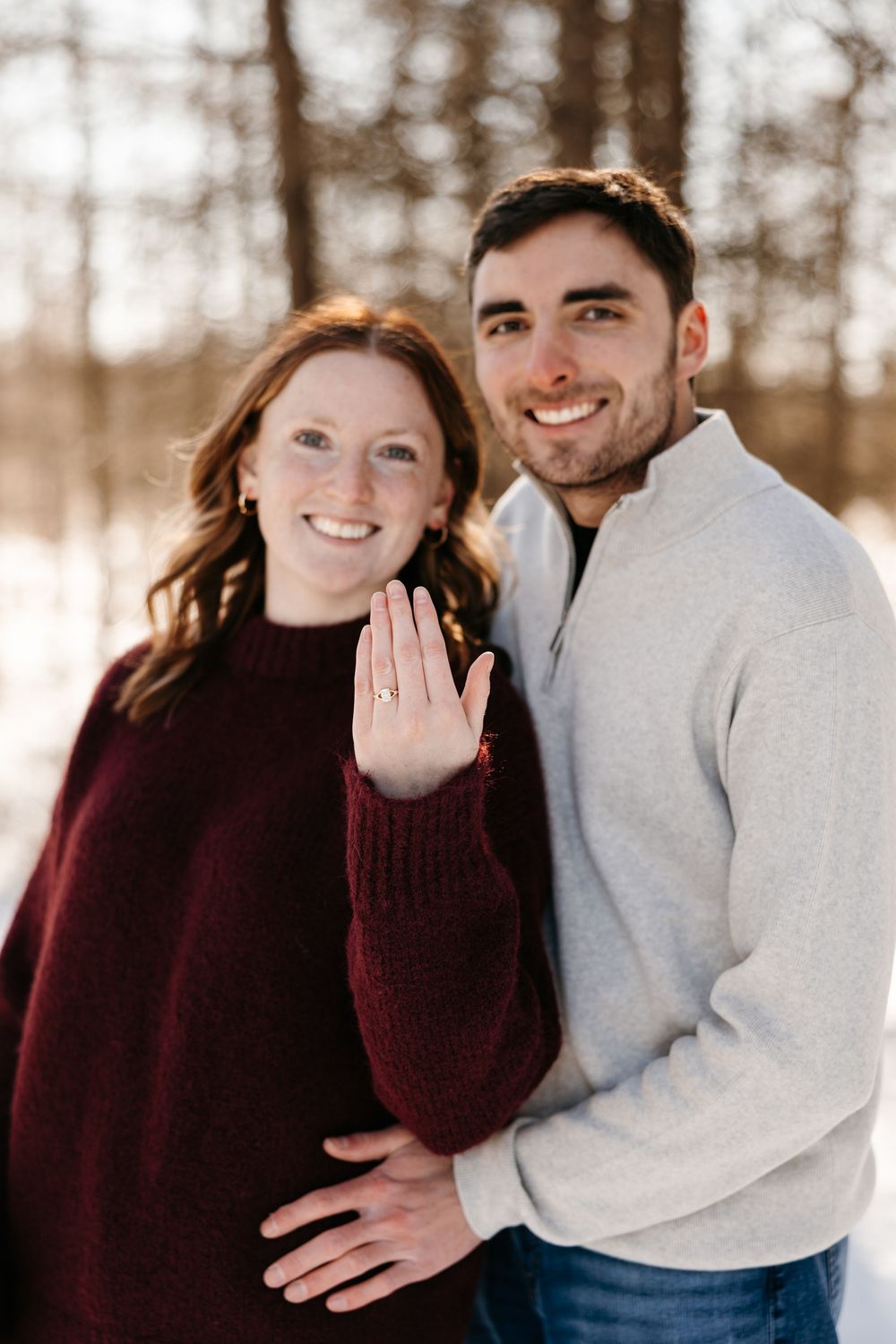 A couple poses together in winter engagement photos wearing burgundy sweater and grey top against snowy backdrop.