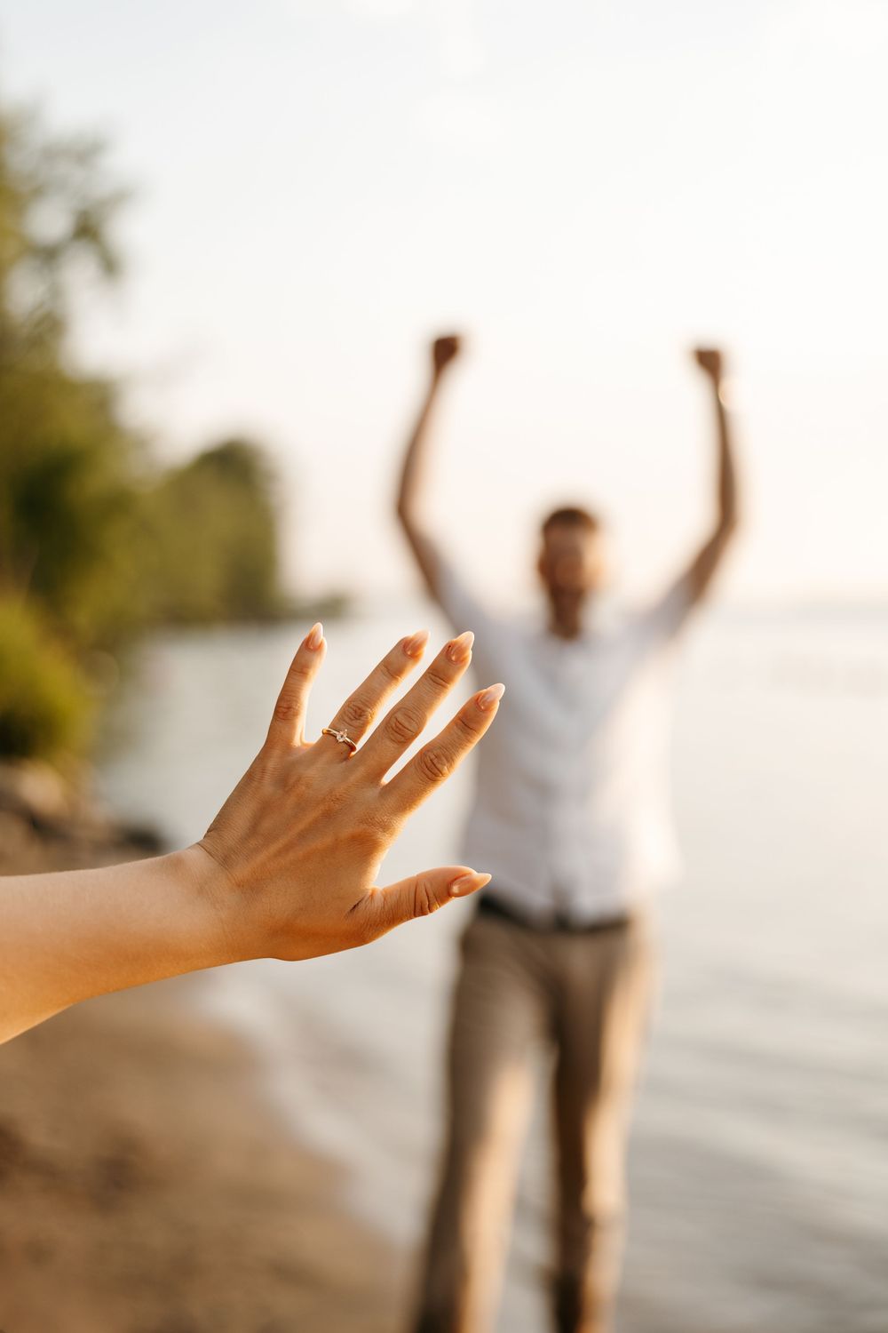 Woman showing engagement ring on beach with excited man celebrating in background near water.