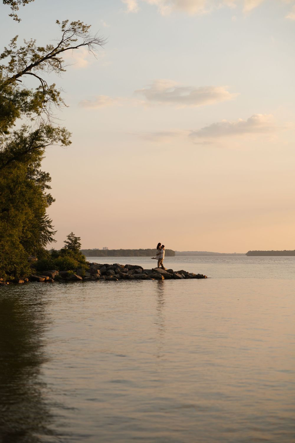 Couple embracing on rocky shoreline at golden hour sunset with calm water and trees.