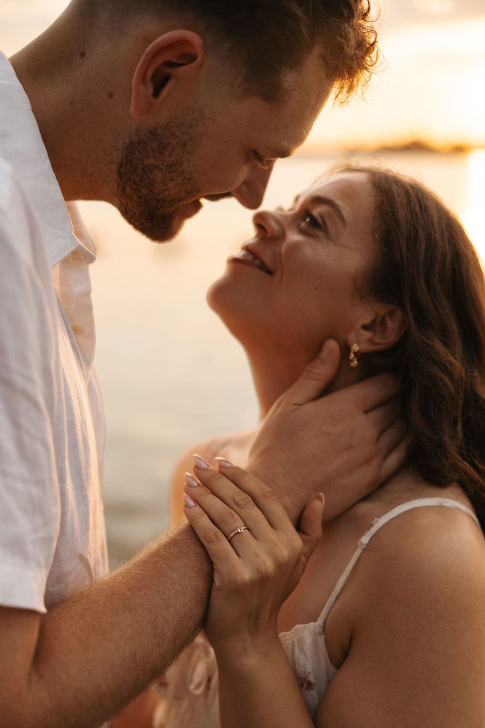 Romantic couple sharing an intimate moment at sunset by the water, woman wearing an engagement ring.