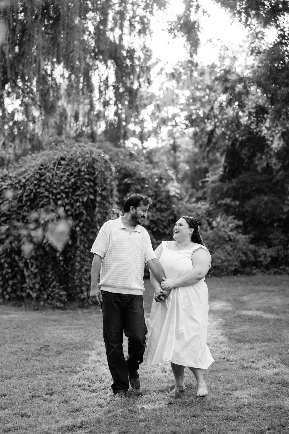 Black and white photo of a couple holding hands and smiling while walking together outdoors in a lush garden.