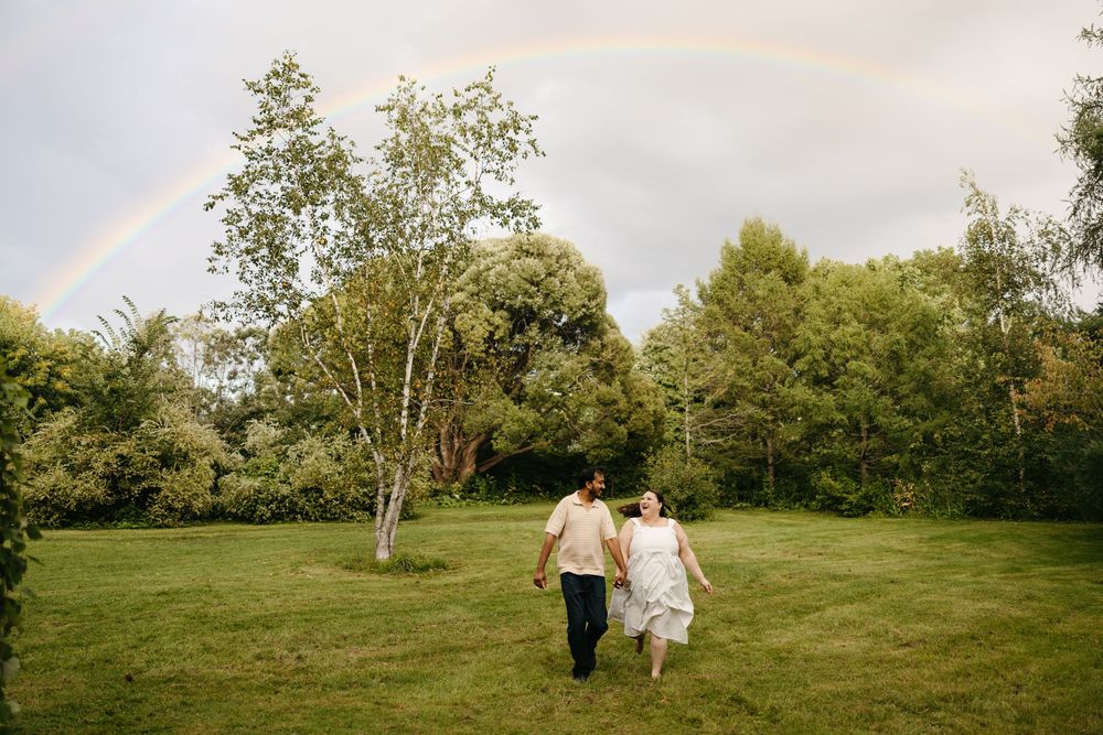 Couple holding hands walking across green lawn with rainbow in sky surrounded by lush trees.
