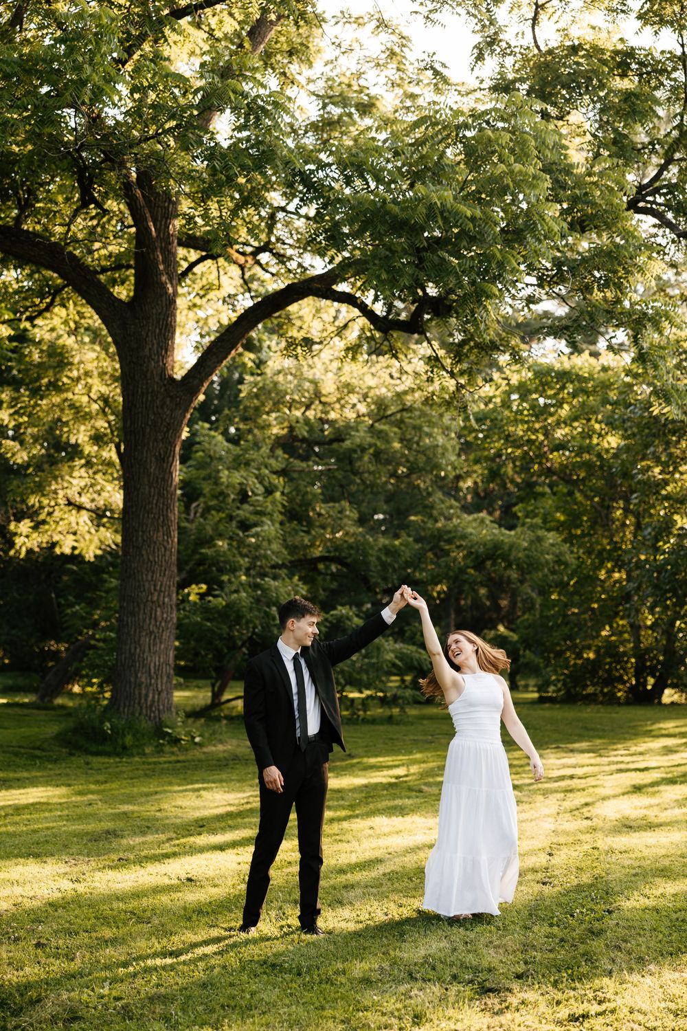 A bride in white dress and groom in black suit dance together in a sunlit park with lush green trees.