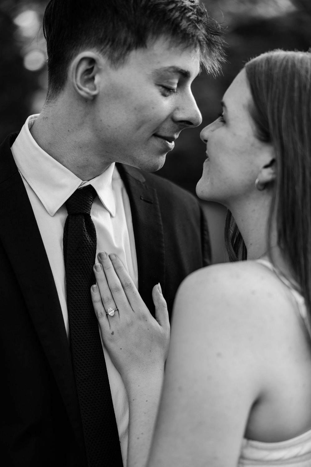 Black and white photo of a couple in formal attire sharing a romantic moment, woman showing engagement ring on mans chest.