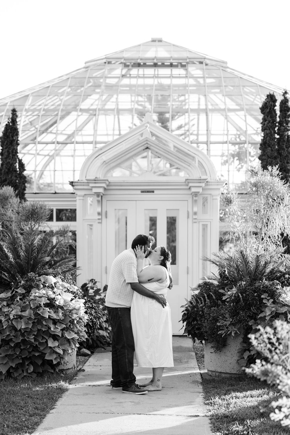 Black and white photo of a couple kissing in front of a stunning glass greenhouse surrounded by lush garden plants.