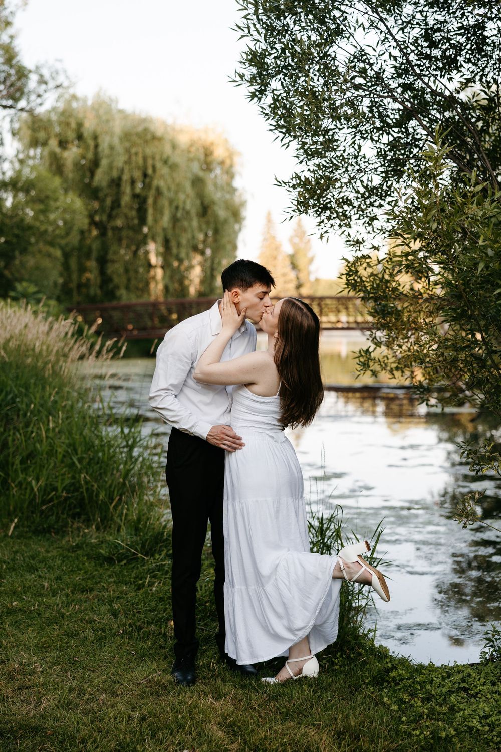 A couple shares a romantic kiss by a serene pond, the bride in a white dress lifting her foot gracefully.