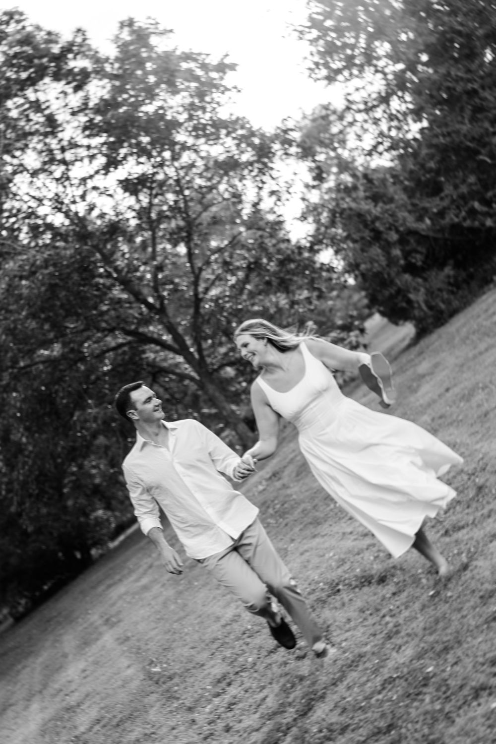Black and white photo of a happy couple holding hands and running through a grassy park with trees in the background.