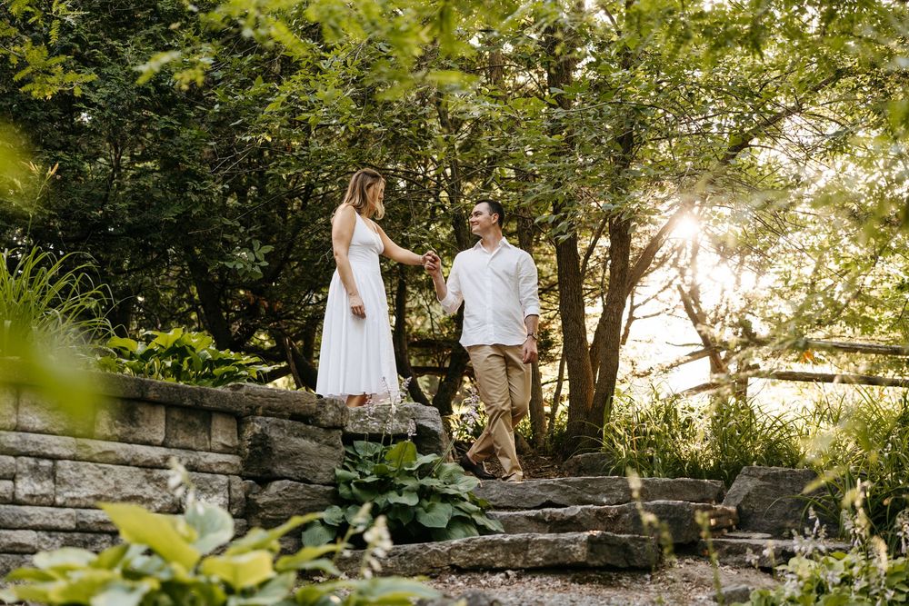 Couple holding hands walking down stone steps in a lush garden at golden hour, woman in white dress.